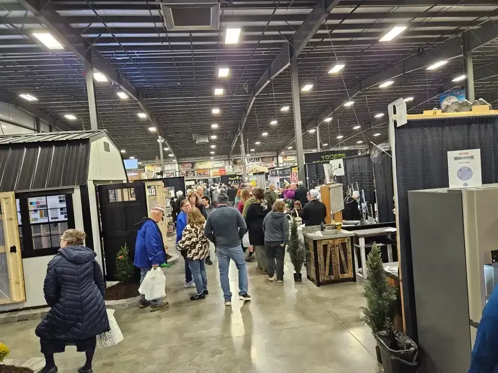 People at an indoor trade show, walking between booths. Many overhead lights, high ceiling.
