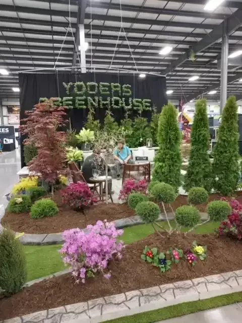 Yoder's Greenhouse display at a trade show. Landscaped garden with colorful plants, trees, and vendor's table.