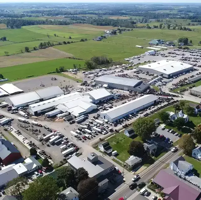 Aerial view of a large outdoor market with various buildings, tents, and many cars parked in a rural setting.