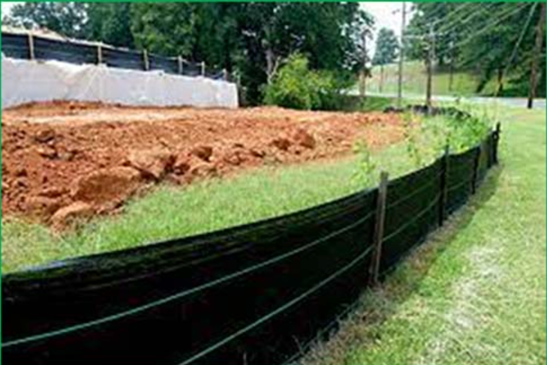 Black silt fence along a grassy edge beside a dirt construction site and trees