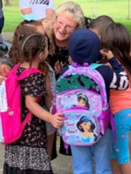 Children with colorful backpacks hug a smiling adult outdoors on a grassy field.