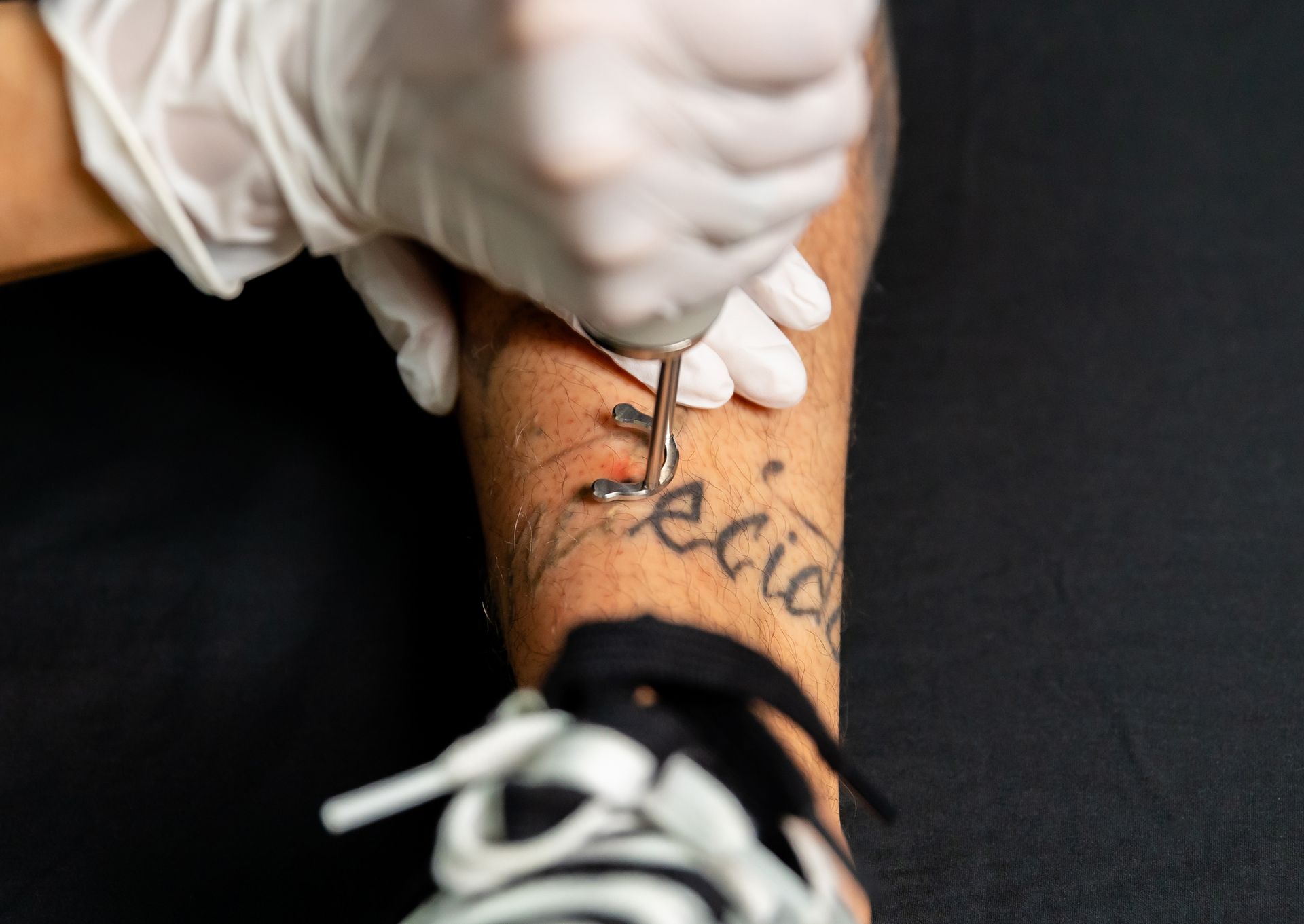 Gloved hands tattooing cursive text on a person’s forearm in a close-up shot