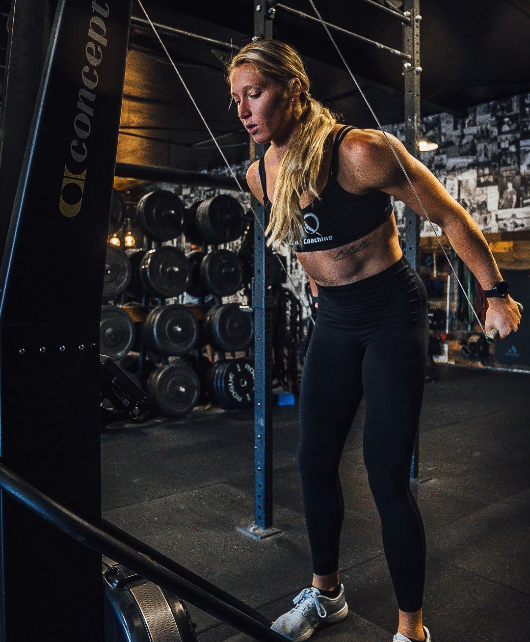 Woman working out with a cable machine in a gym. Blonde hair, wearing black athletic wear, focused expression.