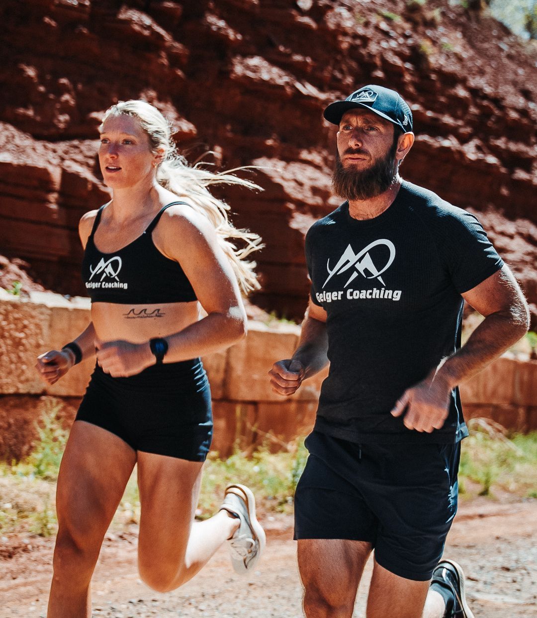 Woman and man running outdoors on a trail. Both wearing athletic clothing, man with a beard and hat.