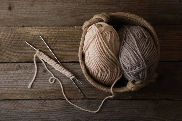 Ferri da maglia con una fila di punti accanto a un cestino contenente due gomitoli di lana su una superficie di legno.