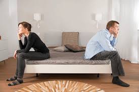Man and a woman frowning, sitting on opposite sides of a bed