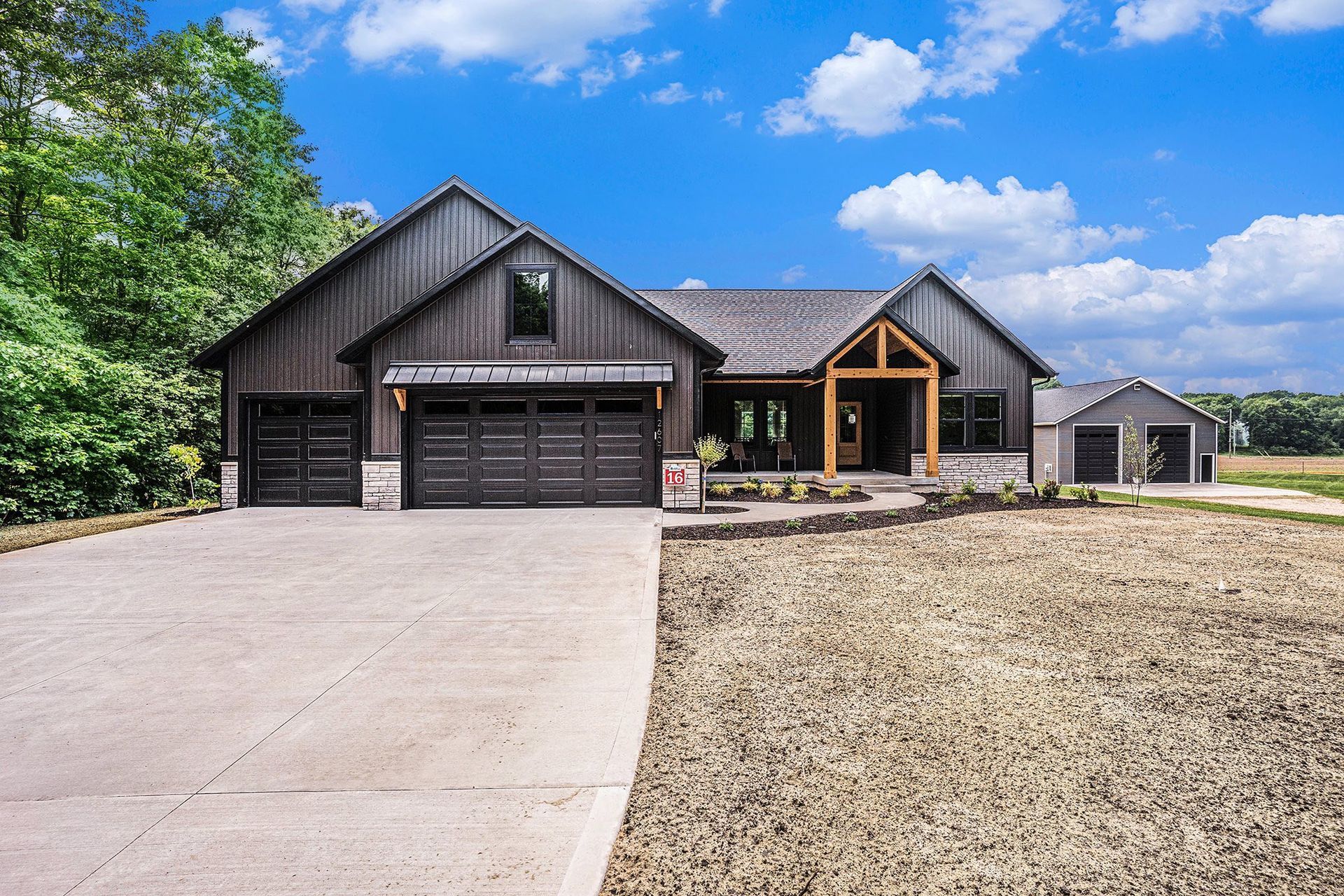 A modern, dark-stained wood home with a large driveway, wood-framed porch, and detached garage on a sunny, grassy lot.