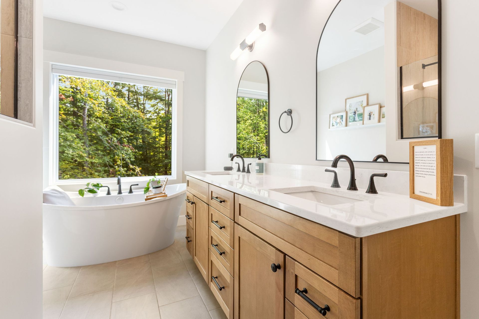 A bright bathroom featuring a white soaking tub by a large window, light wood cabinets, and arched mirrors.