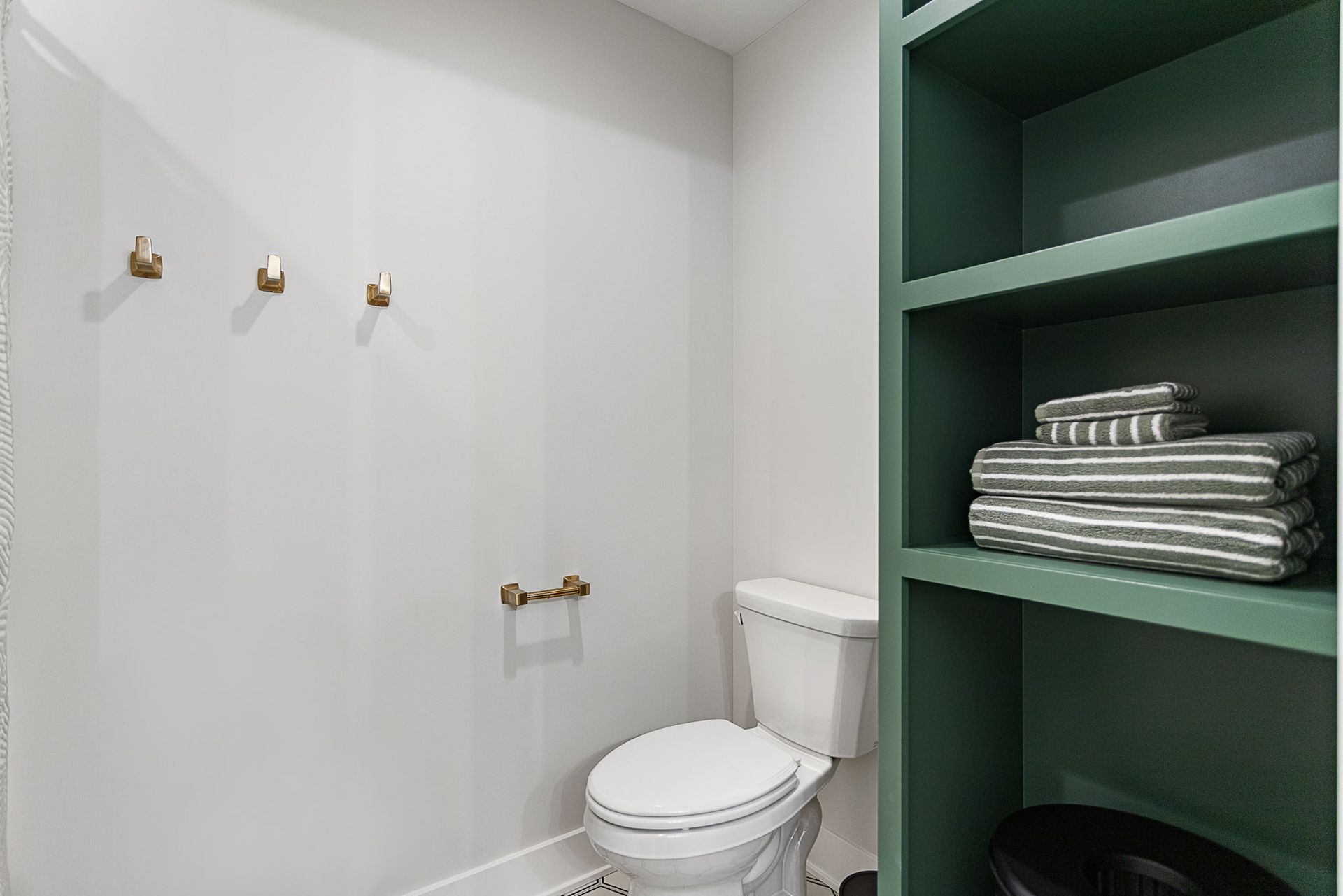 A bathroom with light gray walls, a white toilet, three brass hooks, a brass toilet paper holder, and green open shelves.
