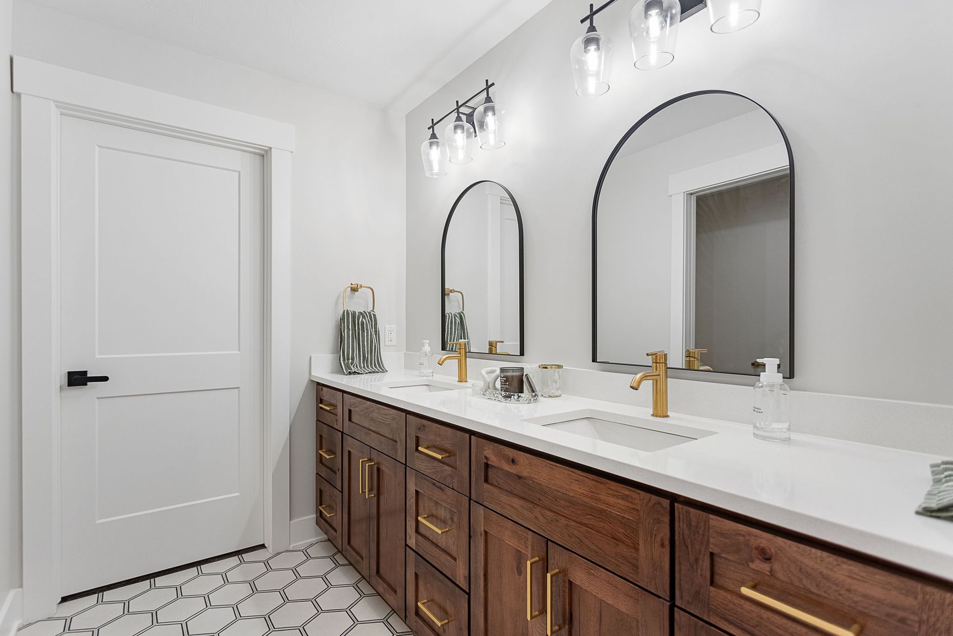 A modern double vanity with dark wood cabinets, white countertops, gold faucets, and arched mirrors in a bright bathroom.