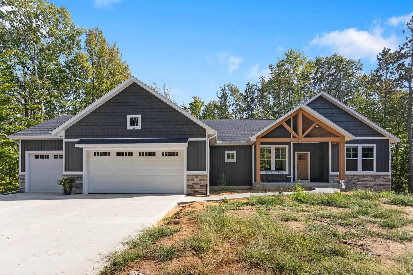 A modern gray house with a two-car garage, stone veneer base, and wood-framed entrance, set against a wooded backdrop.