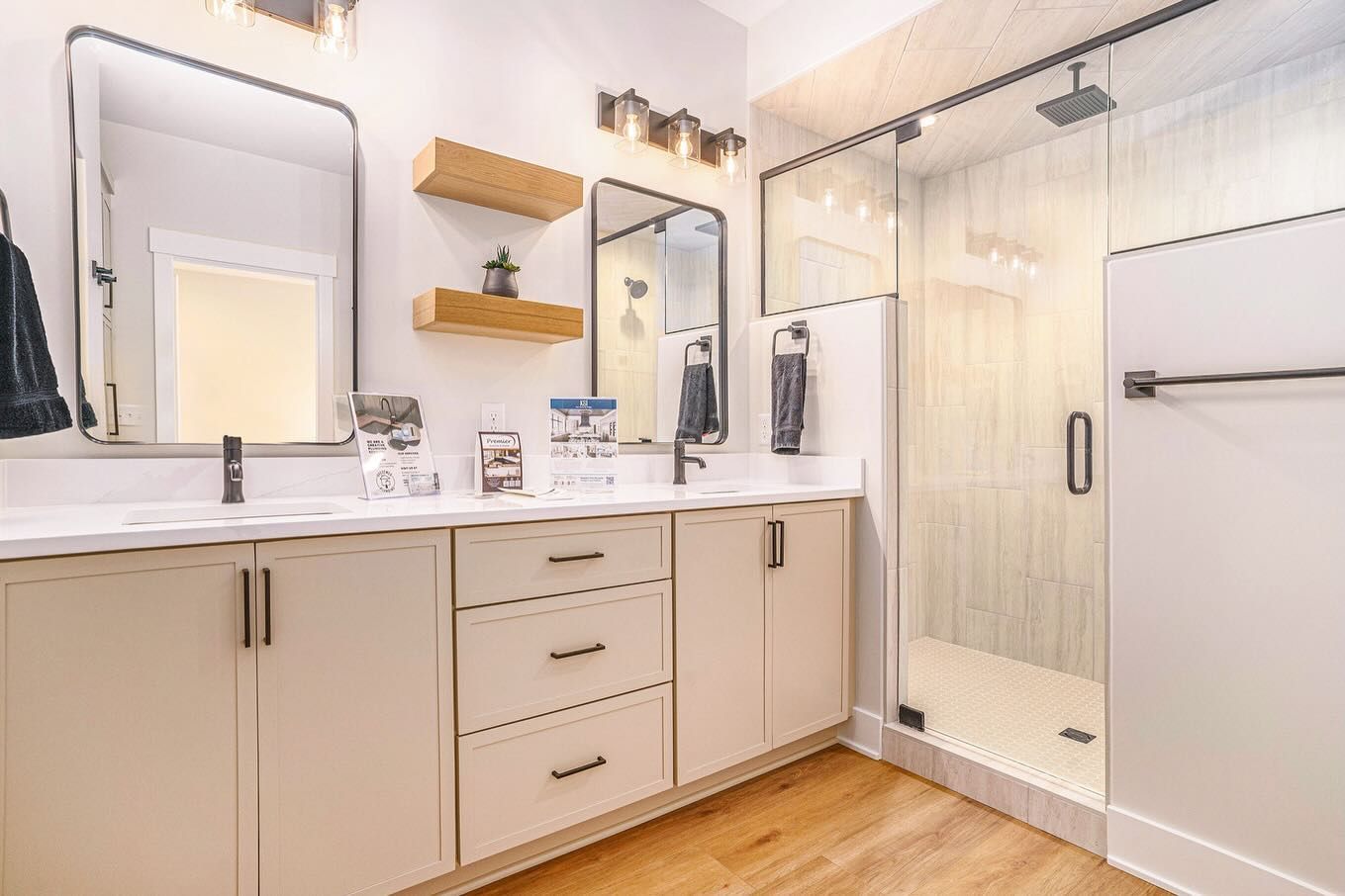 Modern bathroom with a double vanity, light-colored cabinets, two mirrors, wood shelving, and a glass-enclosed shower.