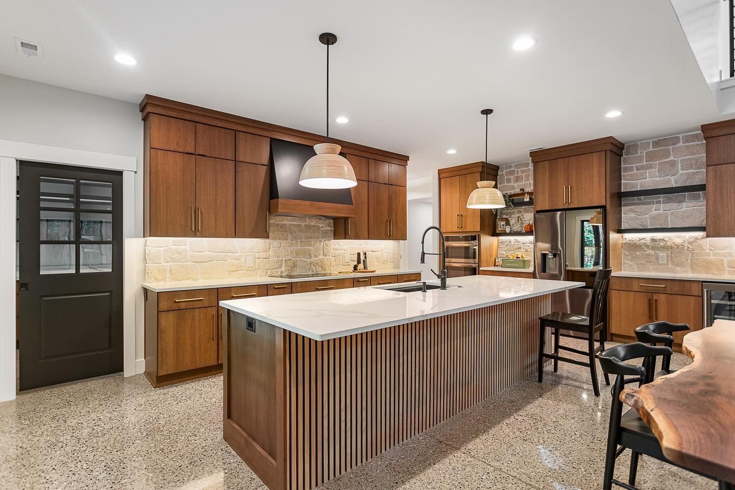A modern kitchen with wood cabinets, a stone backsplash, a large slatted kitchen island, and polished concrete floors.
