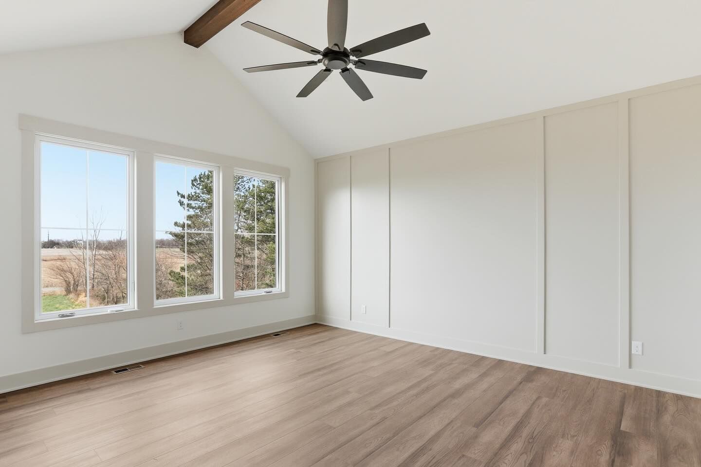 A bright, empty room with light wood floors, a vaulted ceiling, a ceiling fan, and white board-and-batten wall trim.