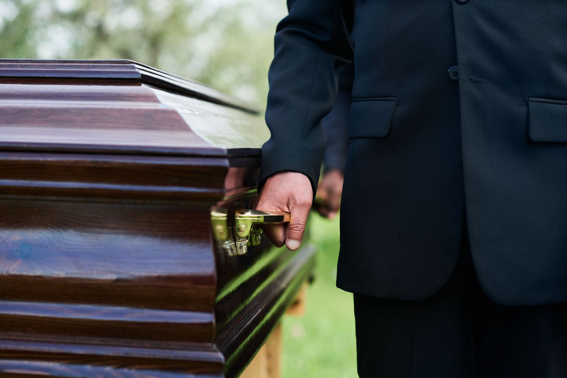 Man in black suit holding a coffin handle, likely at a funeral, outdoors.