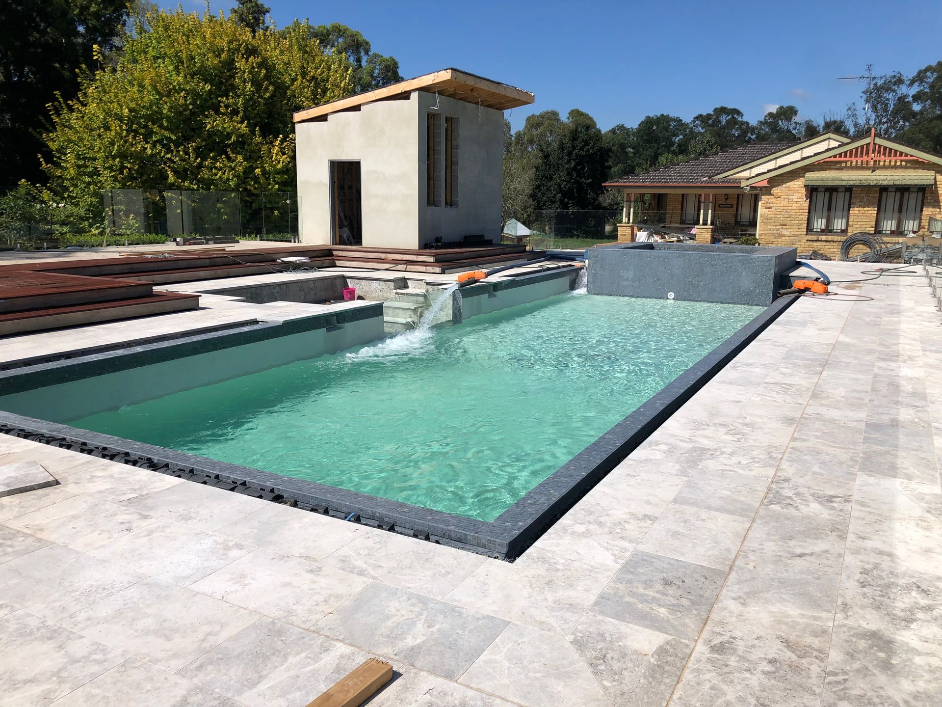 A rectangular swimming pool with light blue water and a gray stone deck, featuring a small building and a house nearby.