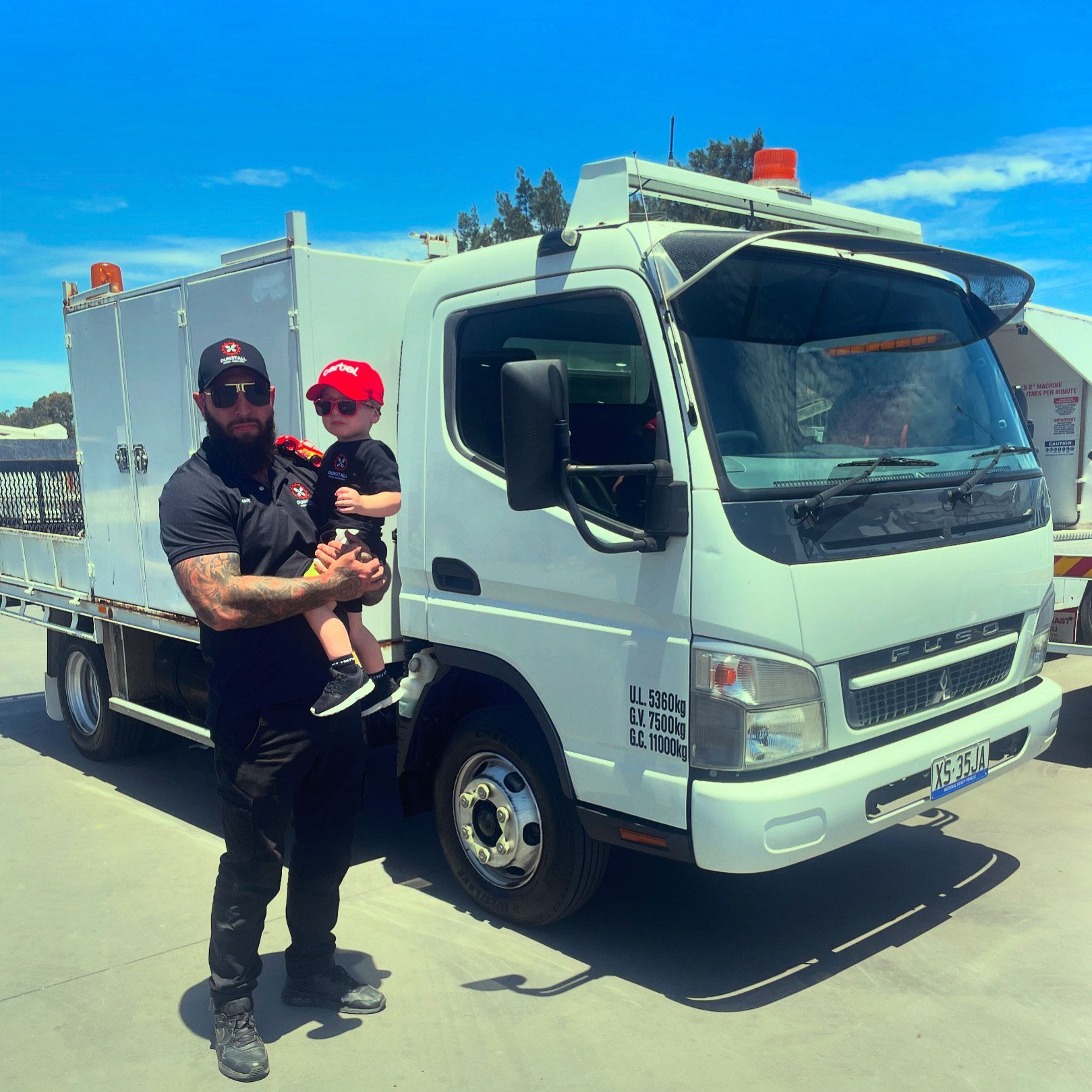 Man holding a child in front of a white utility truck under a bright blue sky.