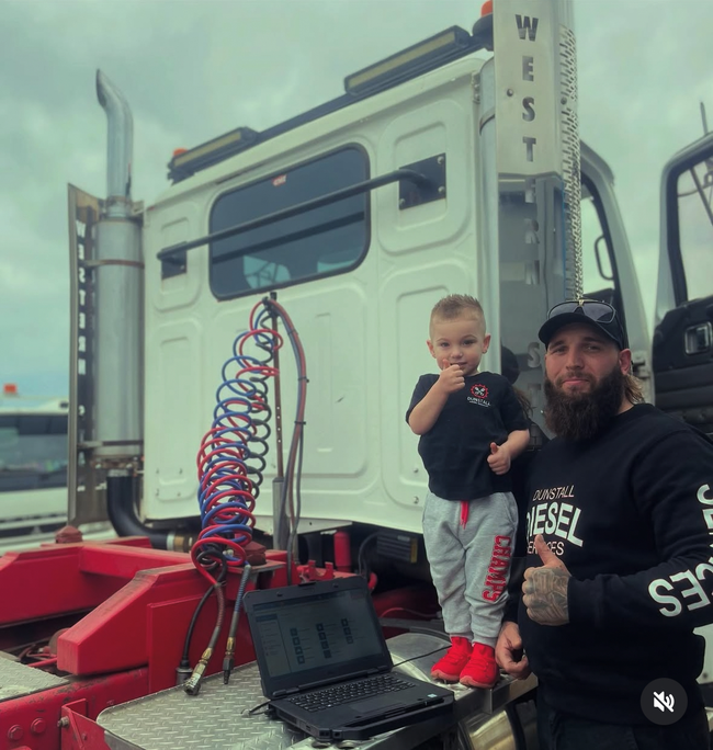 Man and child stand by a white truck, using a laptop. Man with beard, child in red shoes.
