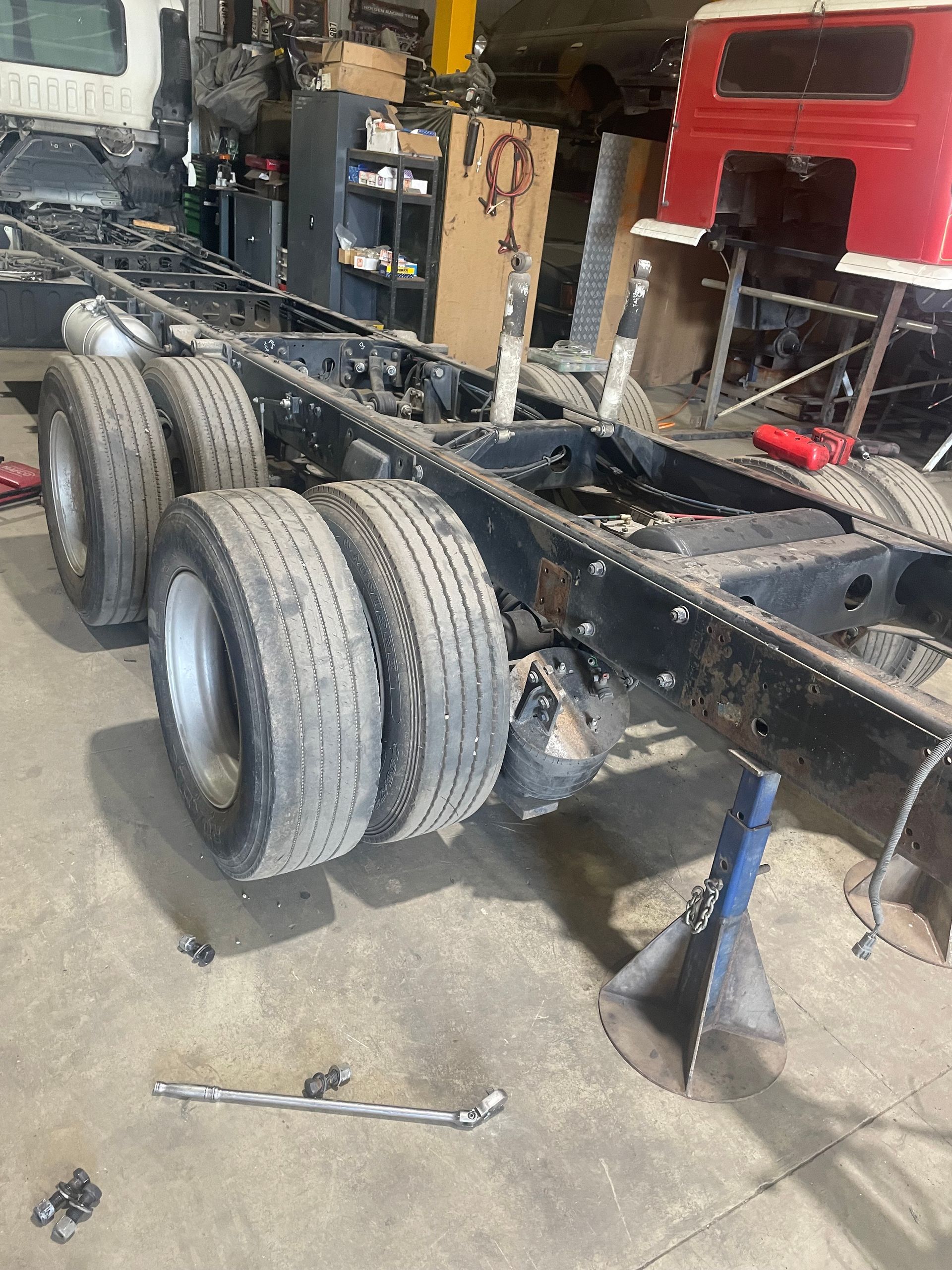 Truck chassis on jack stands in a workshop, tires mounted, tools scattered, red cab in background.