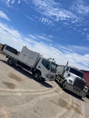 Two work trucks parked on gravel under a blue sky with scattered clouds.