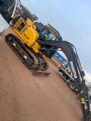 Yellow and black Shantui excavator on a red dirt surface, parked near a truck.