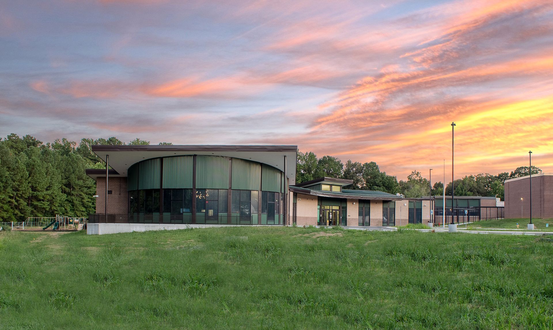 Building with curved facade, tall windows, and green canopy, set against a sunset sky and grassy field.