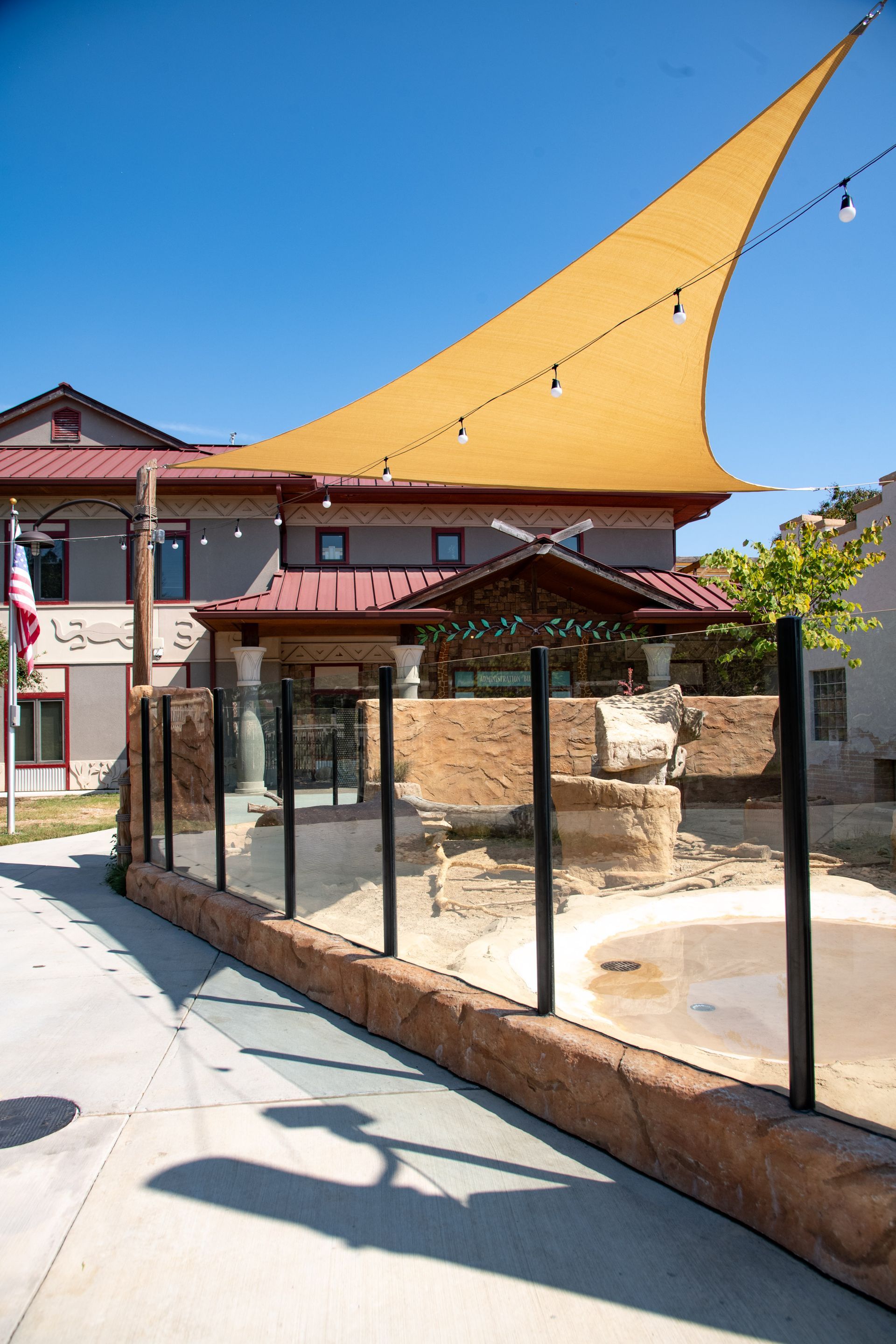 Zoo exhibit with glass barrier and shade, brown and tan facade in background, sunny sky.