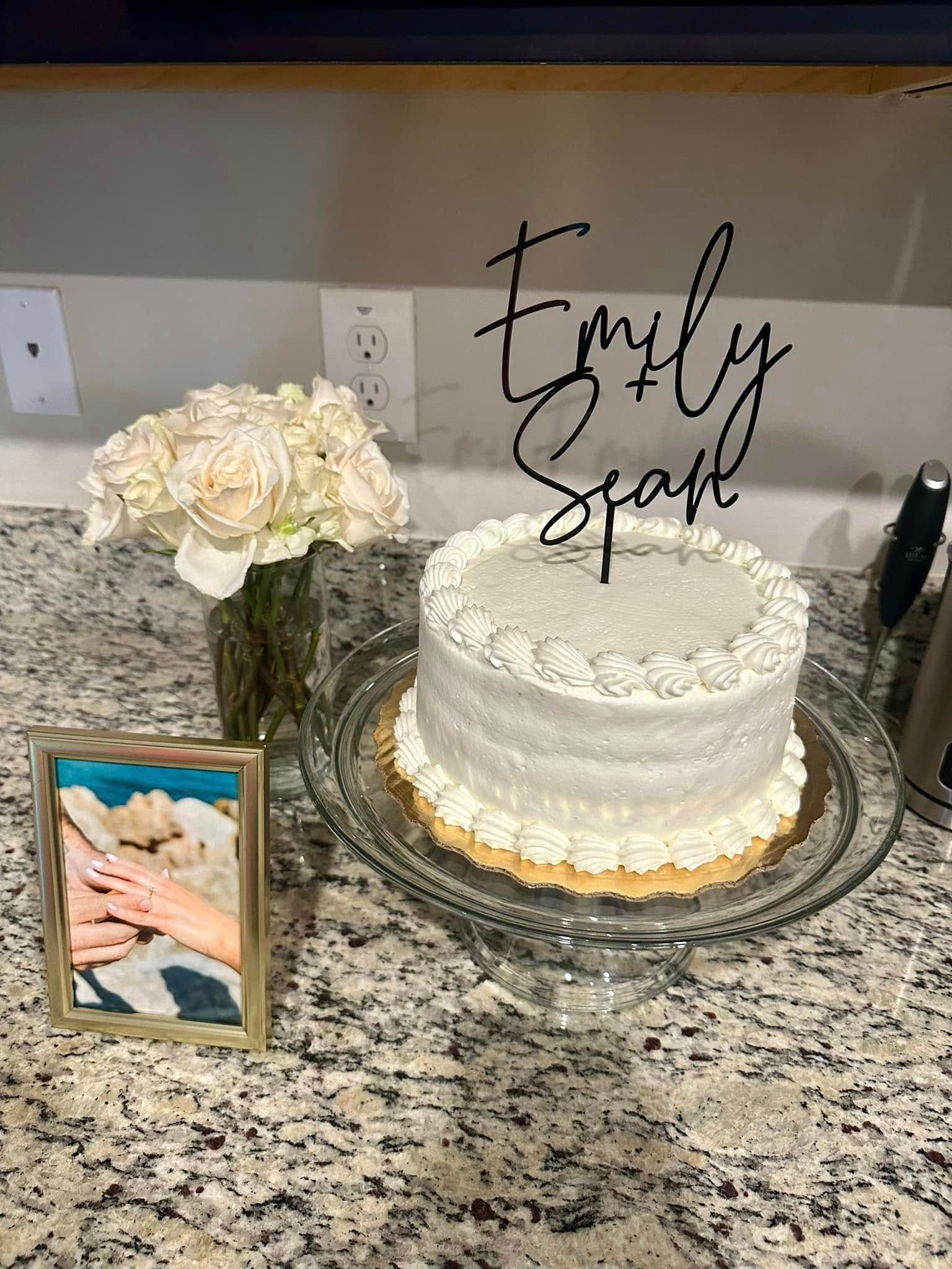 A white cake is sitting on a glass plate on a counter next to a vase of flowers.