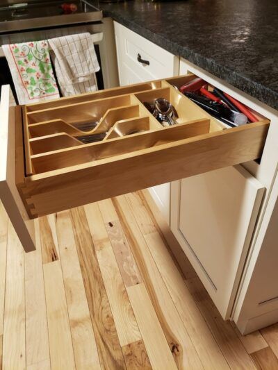 Open kitchen drawer with wooden utensil organizer, next to white cabinets and wood floor.