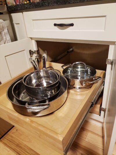A pull-out cabinet drawer filled with stainless steel cookware, in a white kitchen.