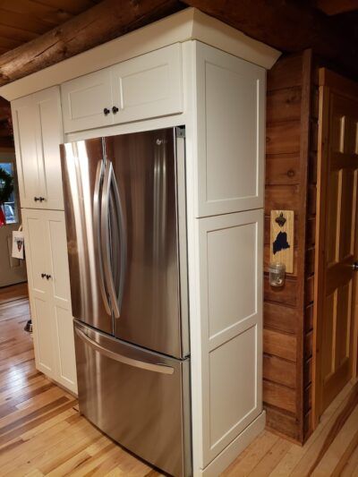 Stainless steel refrigerator built into white cabinetry in a rustic kitchen with wood floors and walls.