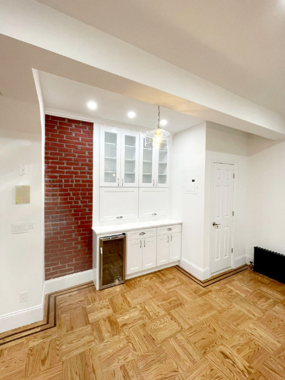 A living room with a brick wall, wooden floor, white cabinets and a refrigerator