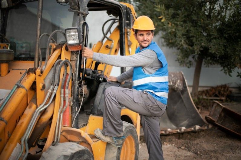 Construction worker in yellow hard hat and blue vest, standing on a yellow backhoe.