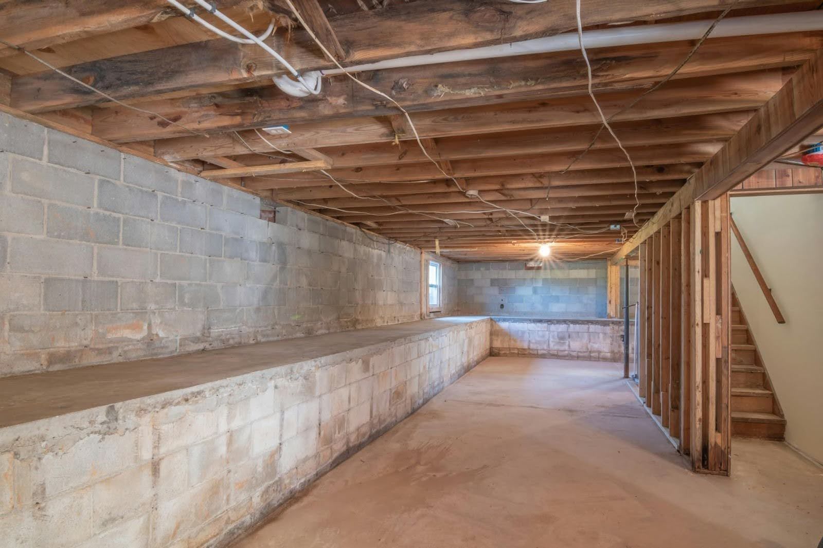 An unfinished basement with exposed wooden ceiling joists, concrete block walls, and a staircase on the right.