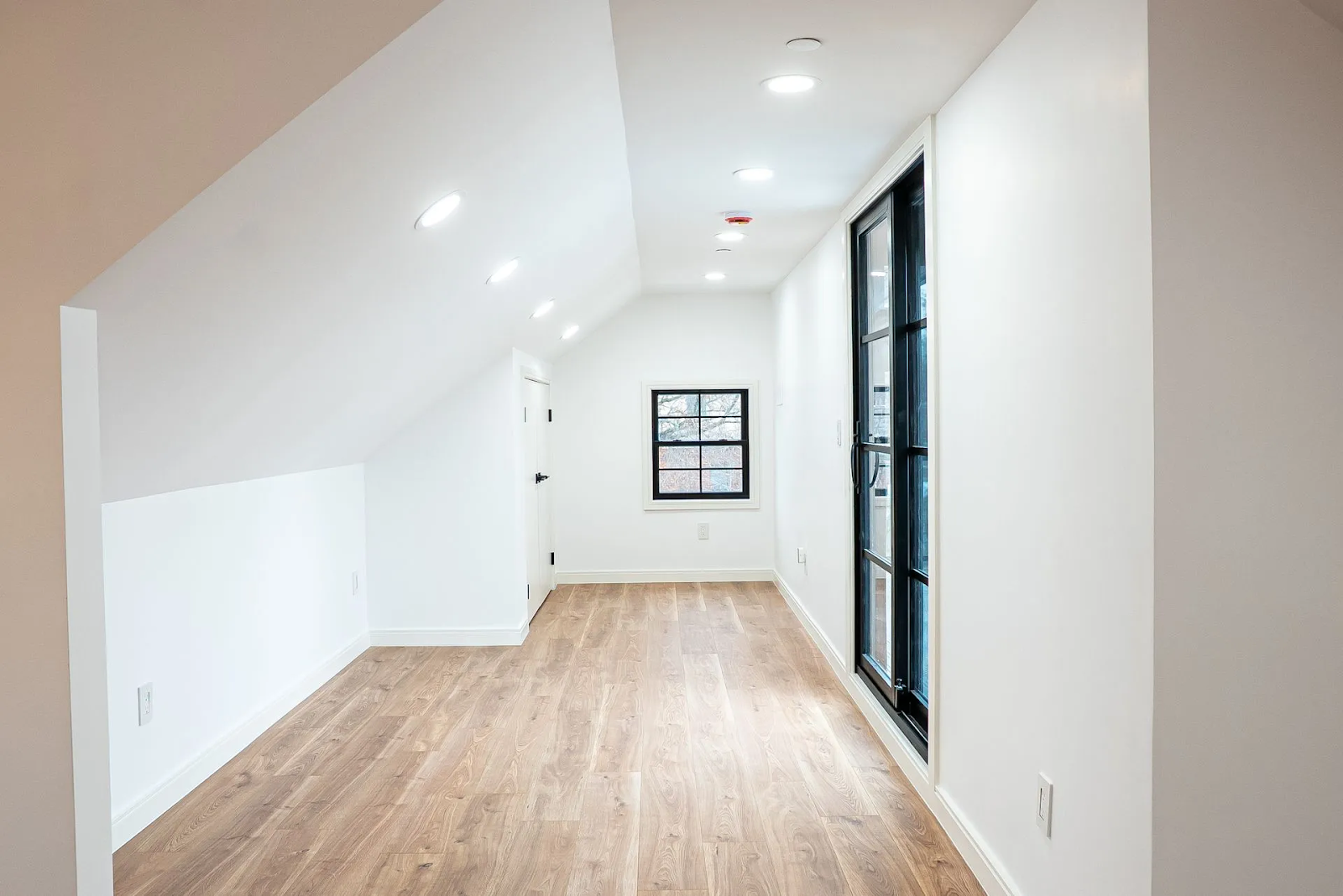 A bright, minimalist attic room with white slanted walls, hardwood floors, a small square window, and black-framed doors.