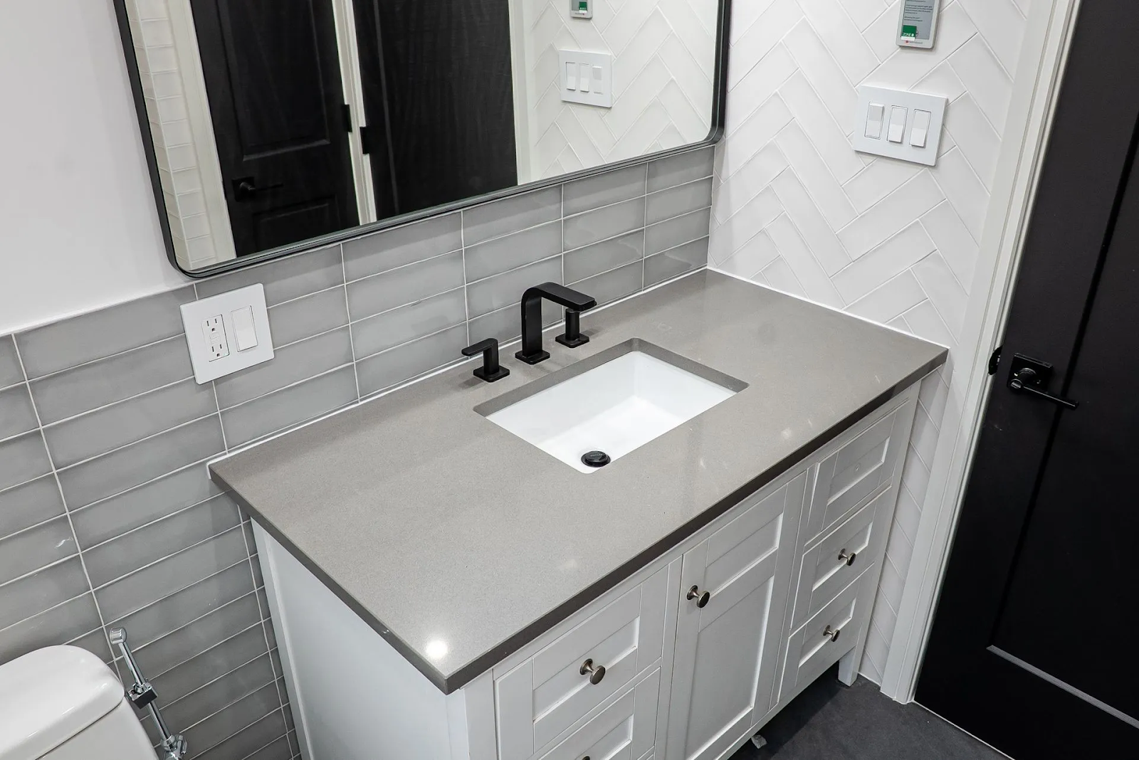 A white bathroom vanity with a grey countertop, black faucet, and rectangular mirror against a grey tiled backsplash.