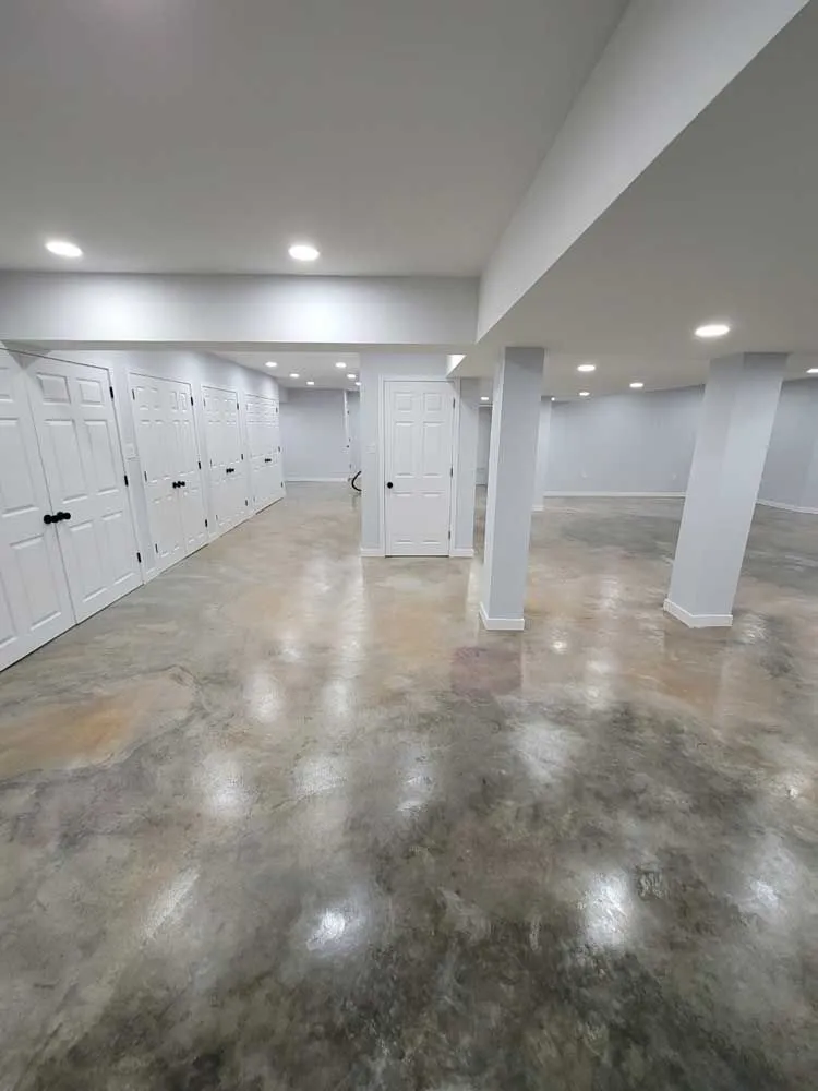 A spacious, unfinished basement with polished concrete floors, white doors along one wall, and two support columns.