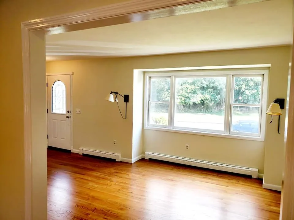 An empty room with polished wood floors, beige walls, a large window, and wall-mounted light fixtures.