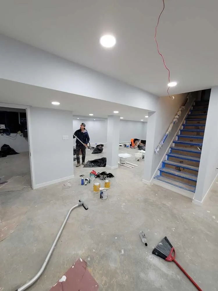 Two workers in a basement renovation, with unfinished concrete floors, newly painted walls, and stairs under construction.