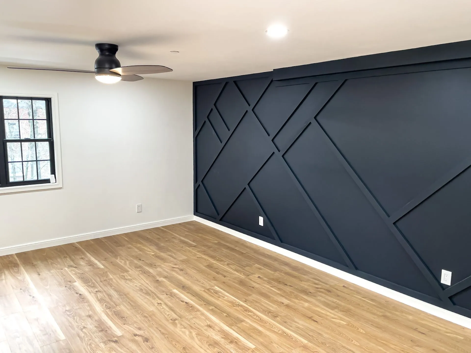 A room with light wood floors, white walls, a ceiling fan, and a large navy blue accent wall with geometric wood molding.