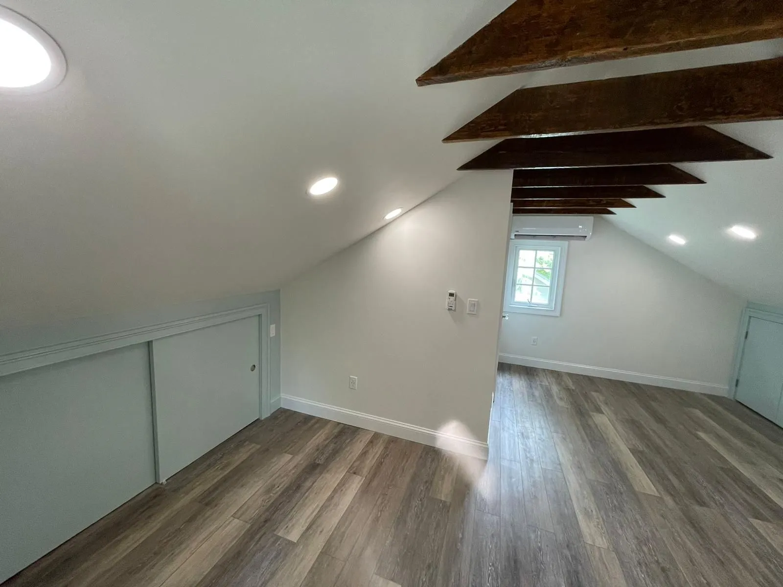 Attic room with sloped white walls, dark exposed wood ceiling beams, light wood-grain flooring, and recessed lighting.