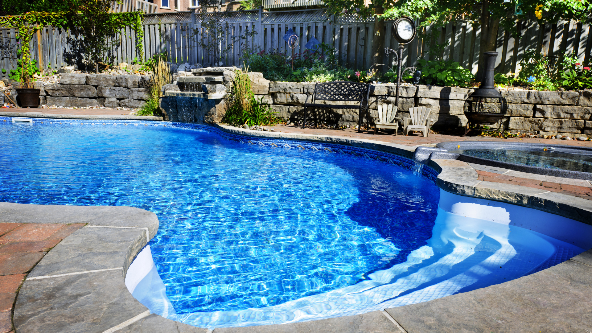 A person cleans a backyard pool using a long-handled pole, with a coiled blue hose lying on the artificial grass nearby.