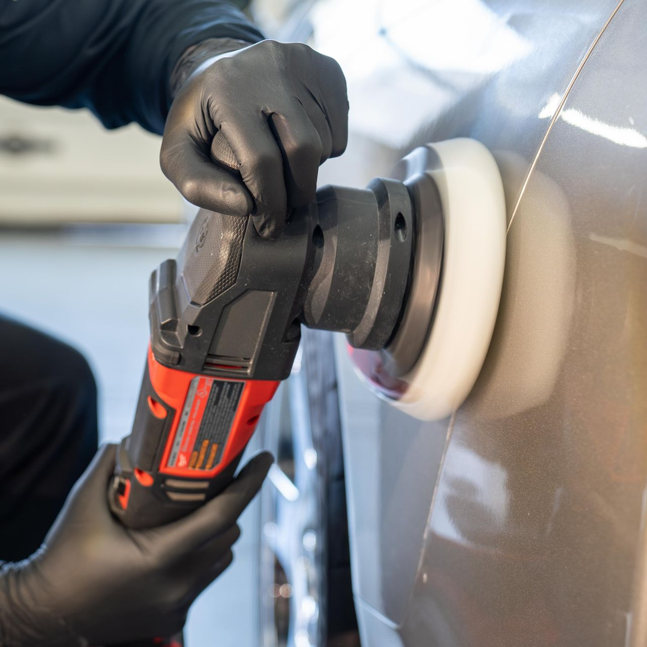 Person wearing black gloves polishing car with an electric buffer in a garage.