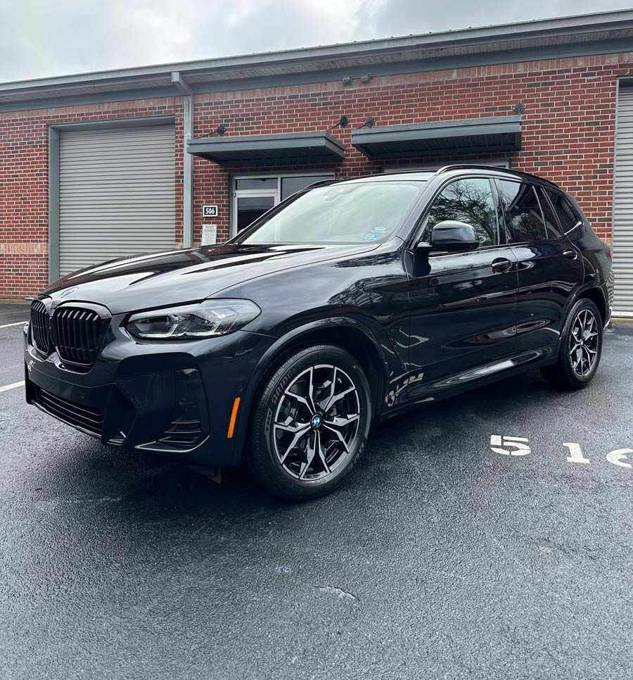 Black BMW X3 parked in front of a brick building with garage doors. Gray asphalt in the foreground.