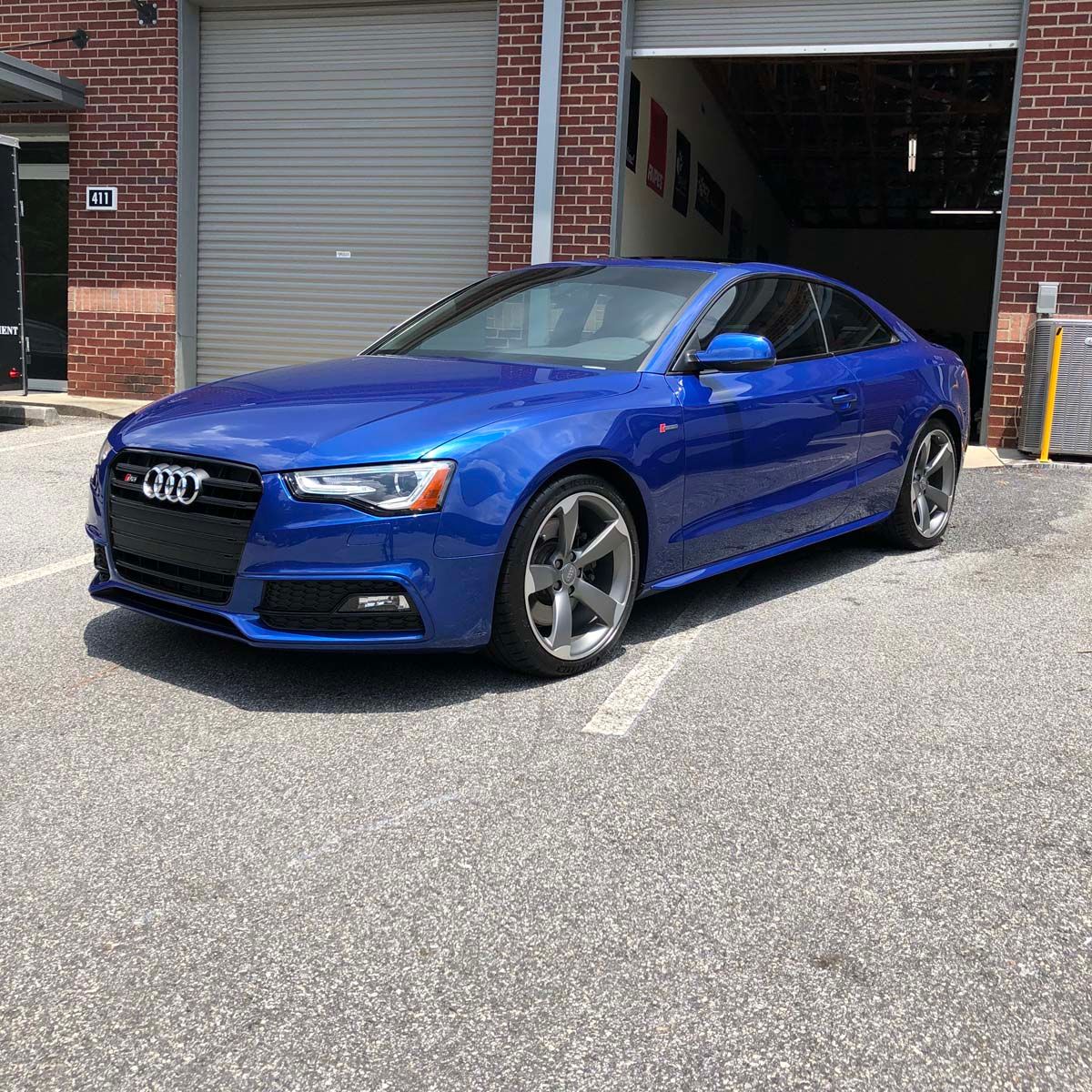 Blue Audi A5 coupe parked in front of a garage, on a paved surface.