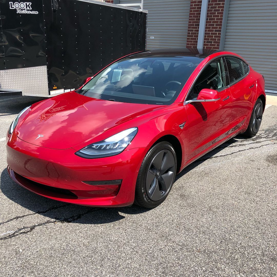 Red Tesla Model 3 car parked on pavement next to a building.