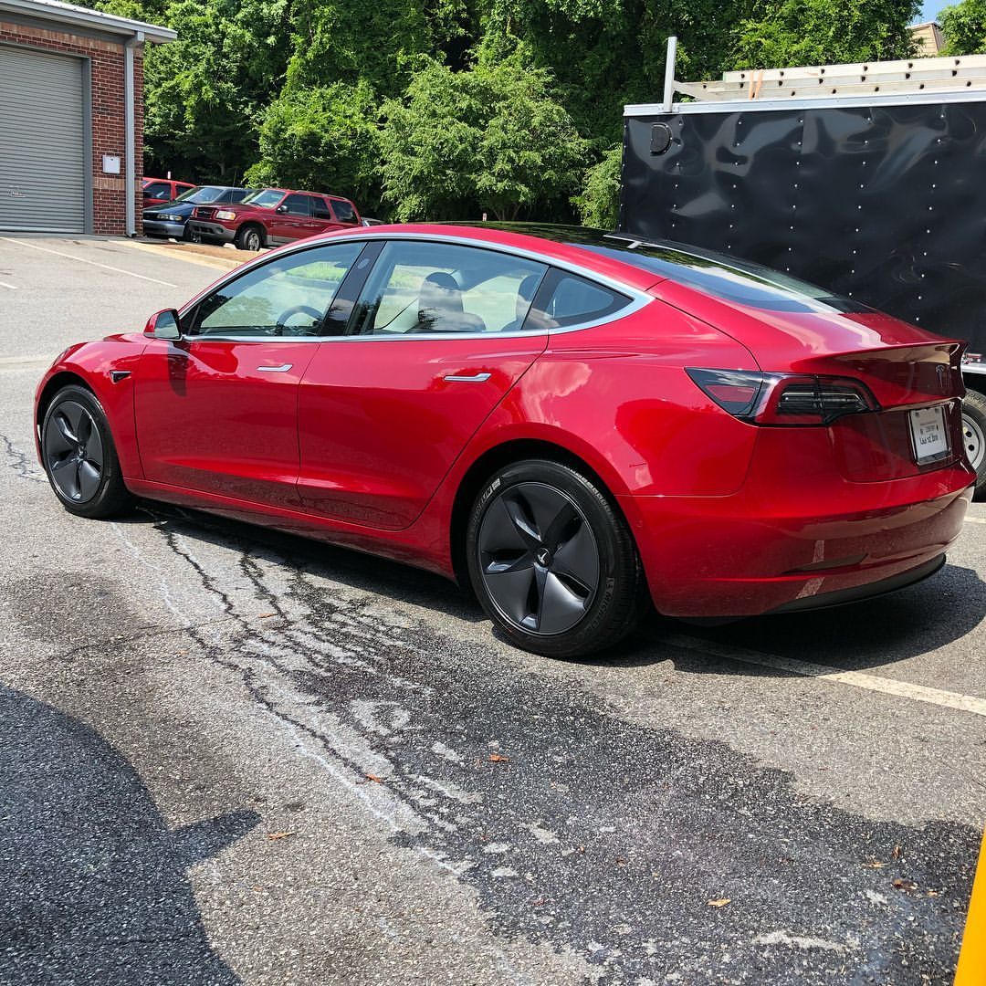 Red Tesla Model 3 car parked on pavement. Black roof and wheels.