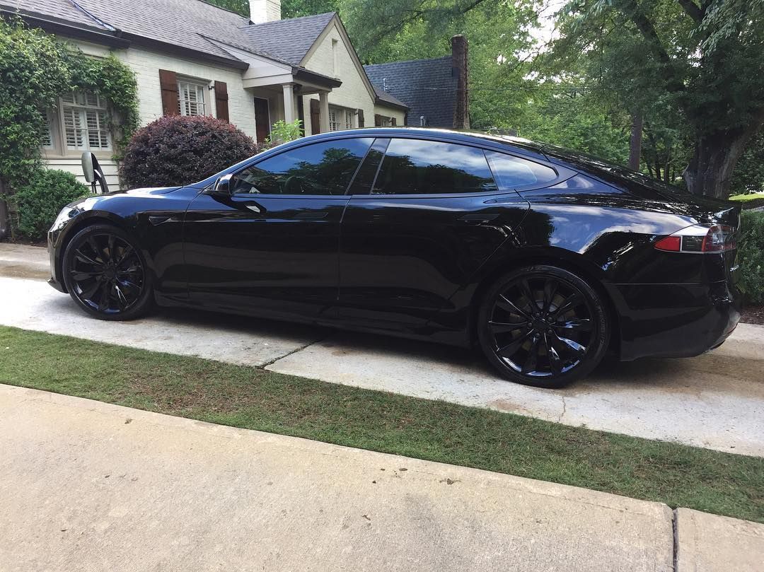 Black Tesla sedan parked in front of a house, with tinted windows and black wheels.