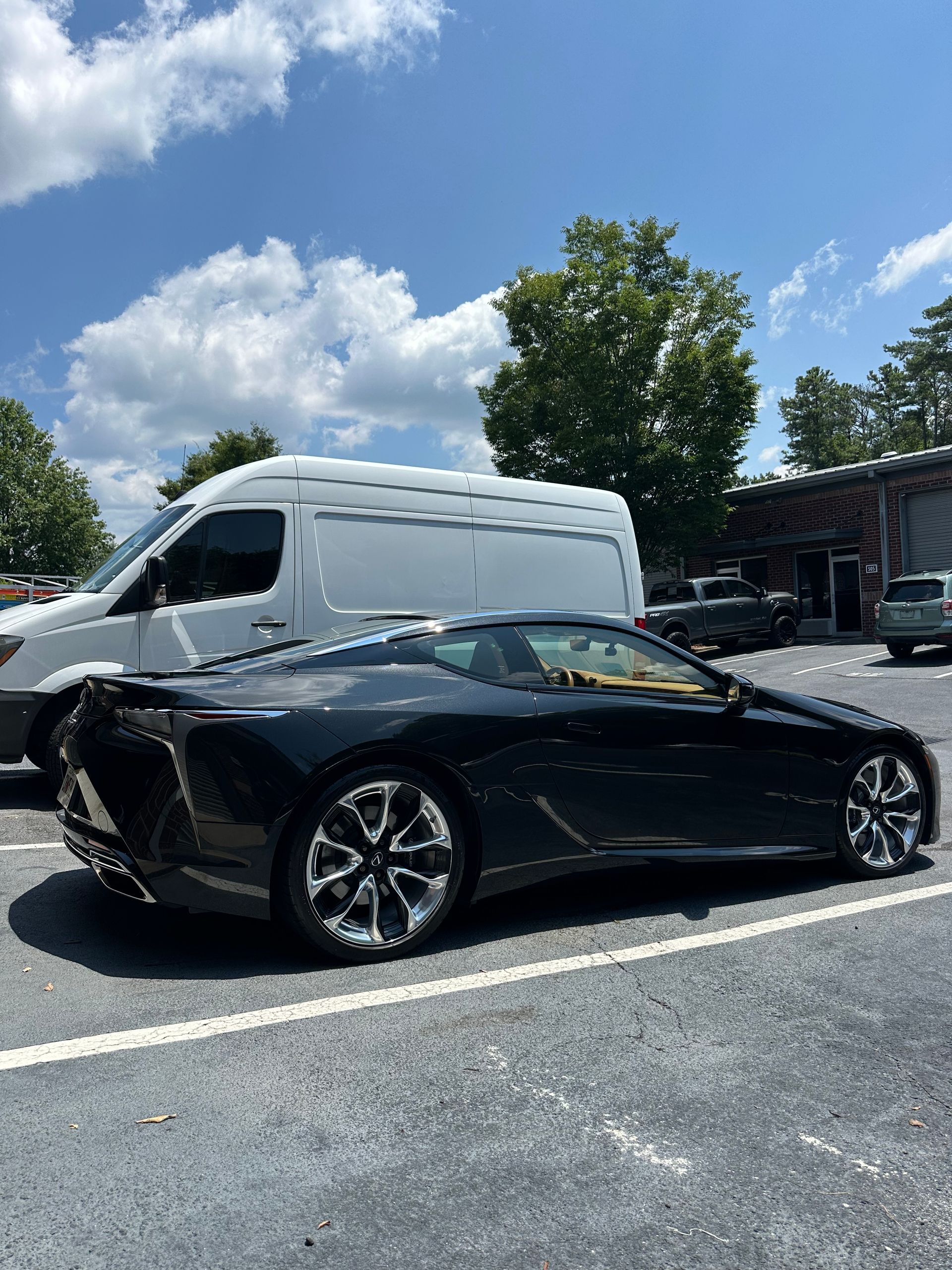 Black Lexus coupe parked next to a white van in a parking lot on a sunny day.