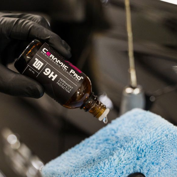 Black-gloved hand applying ceramic coating from a brown bottle onto a blue microfiber cloth, near a car.